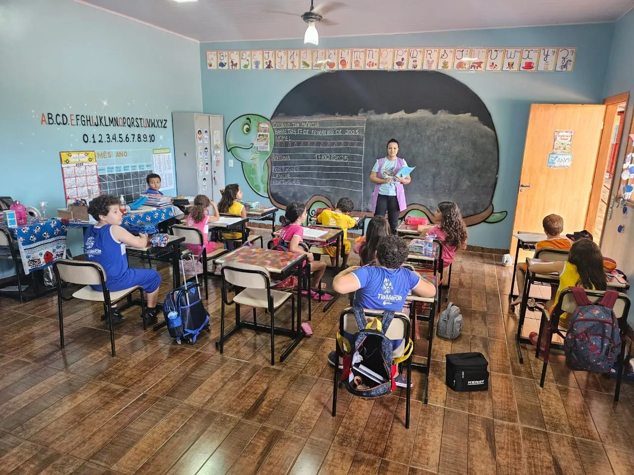 Alunos de uniforme azul em aula, com a lousa em formato de tartaruga ao fundo