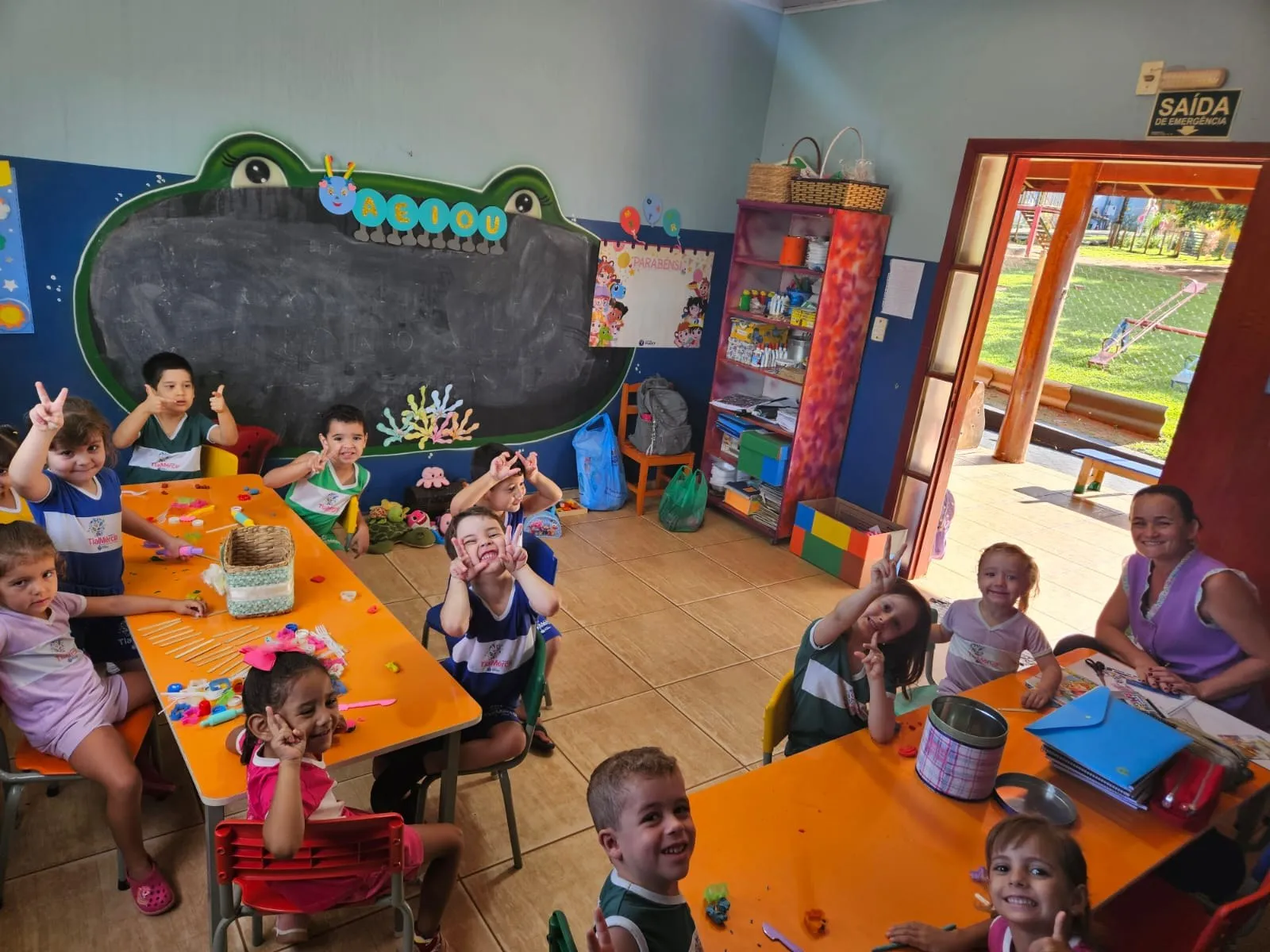 Crianças sorrindo e fazendo sinal de “V” com os dedos em volta das mesas laranja de uma sala de aula do Colégio Tia Mércia
