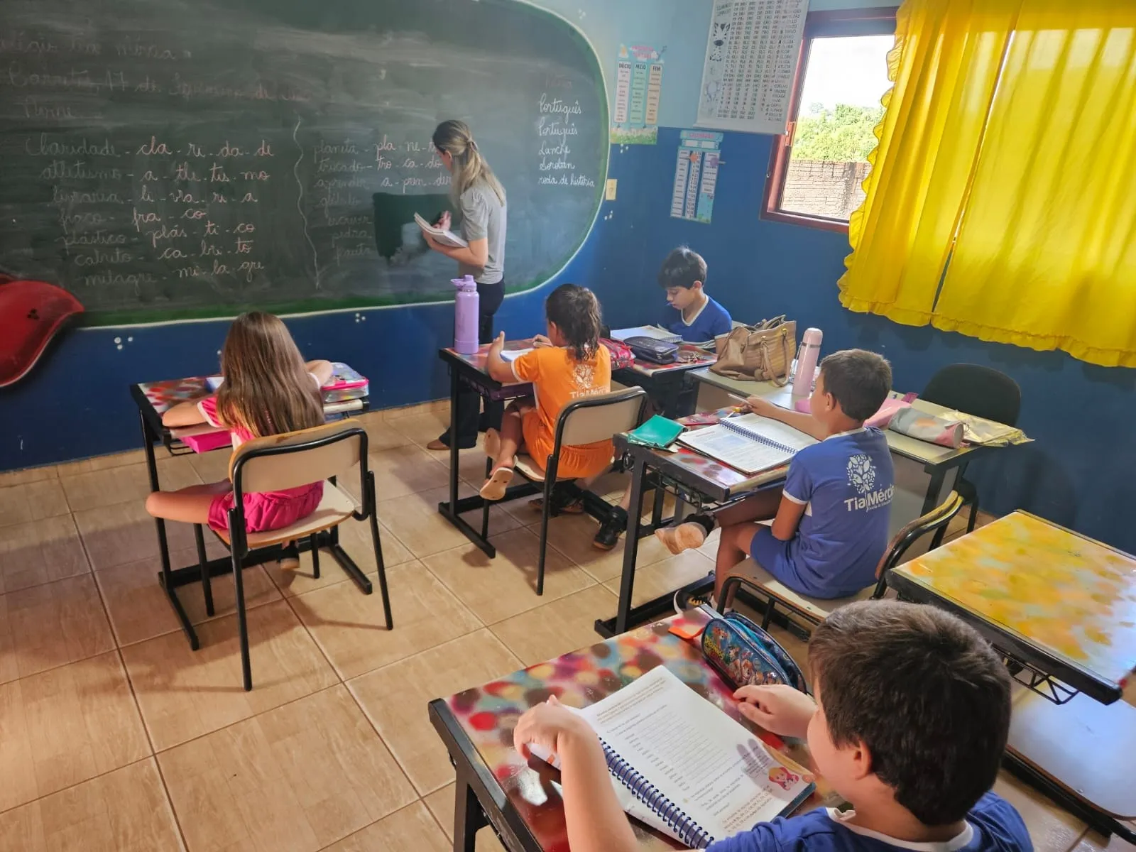 Professora escrevendo na lousa enquanto os alunos copiam nos cadernos