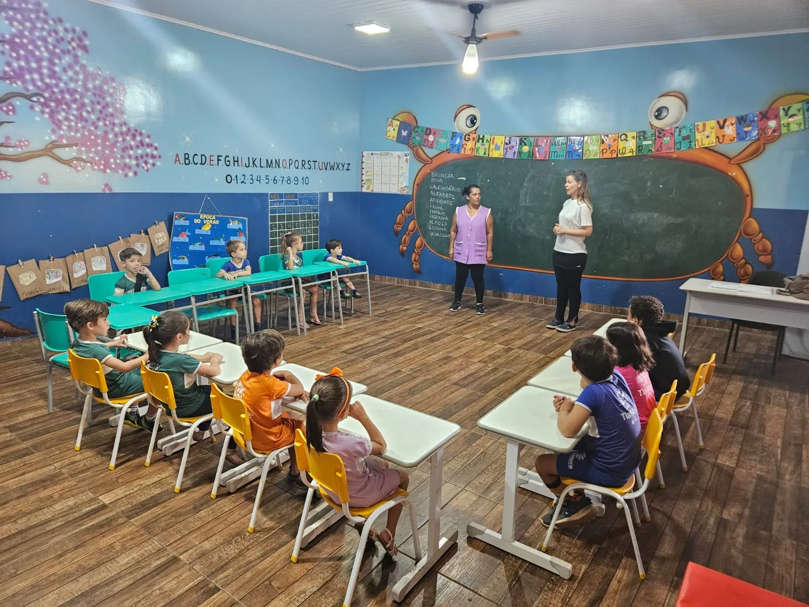 Sala de aula ampla e colorida, com lousa em formato de caranguejo, mural de árvore florida e alunos em suas carteiras