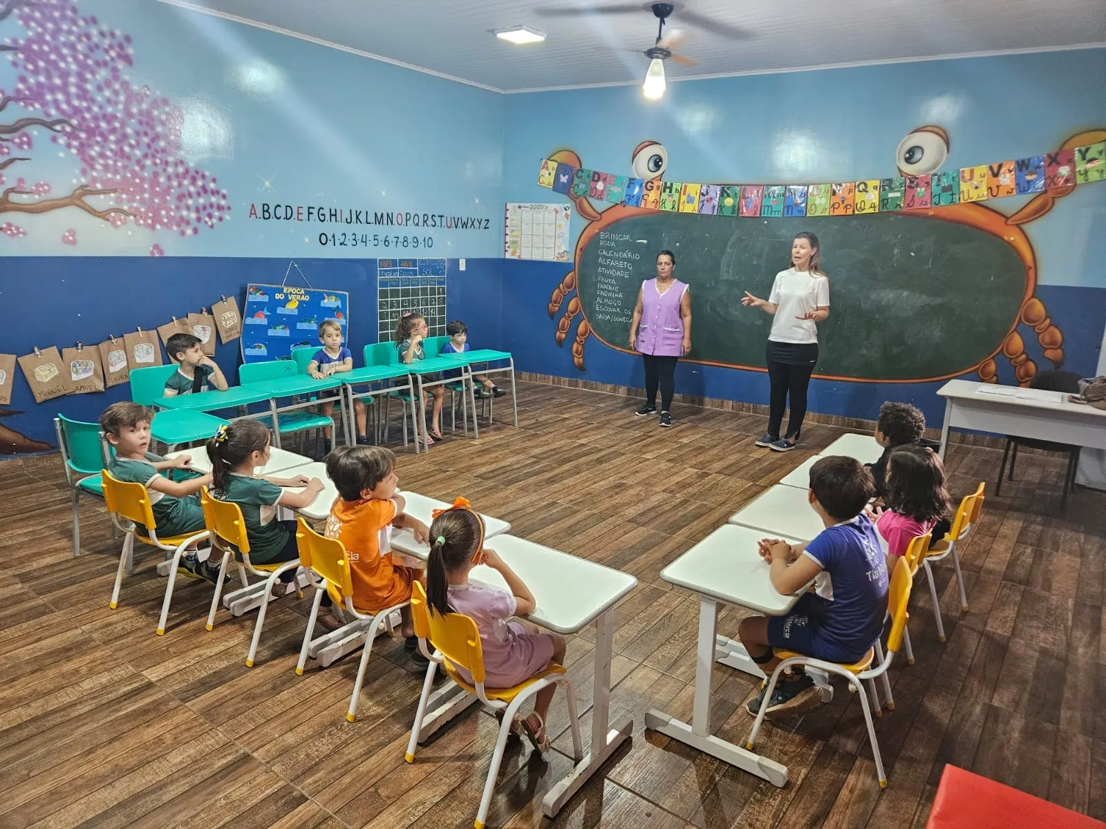 Alunos sentados em carteiras coloridas diante da lousa em formato de caranguejo