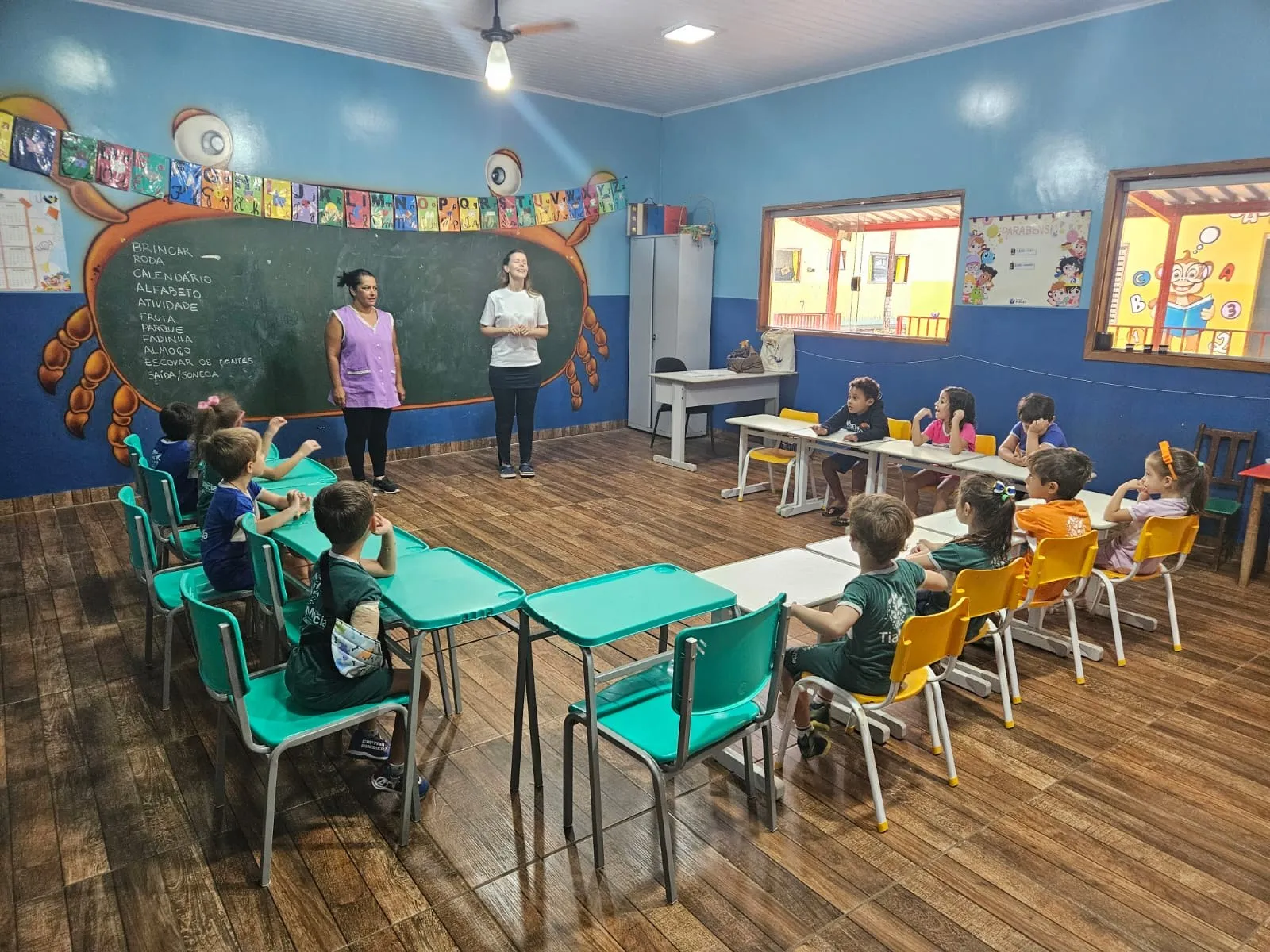 Duas professoras conversando com a turma, em sala com carteiras verde-água