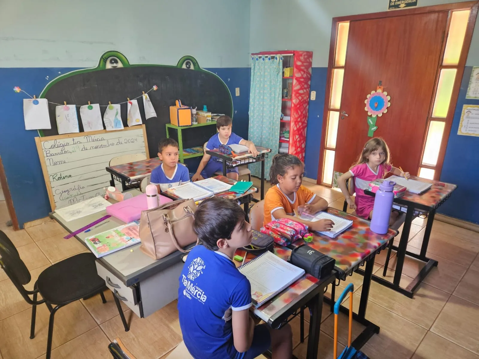 Alunos fazendo atividades em suas carteiras, com trabalhos pendurados na parede