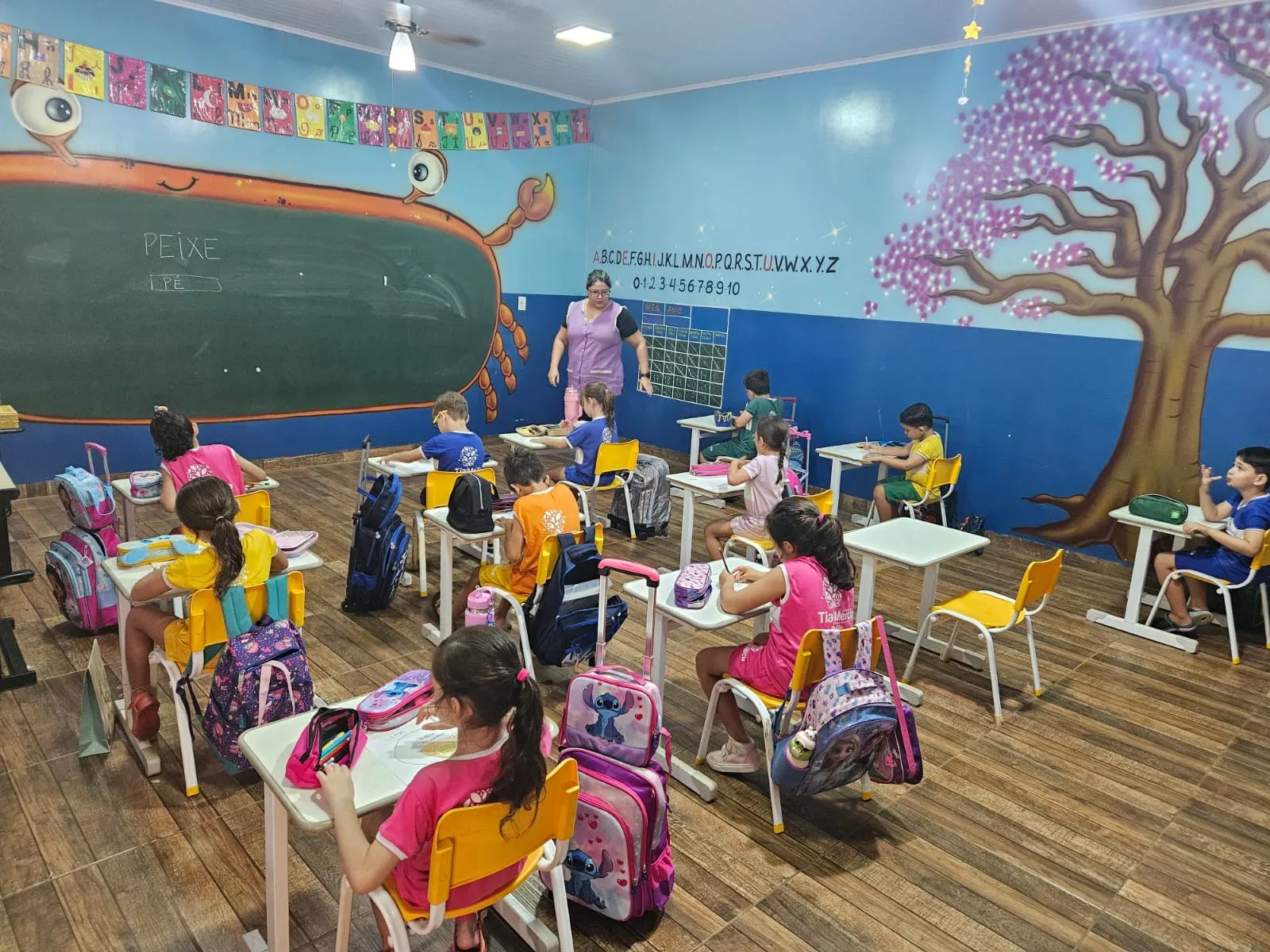 Alunos com mochilas coloridas, em sala com mural de árvore florida